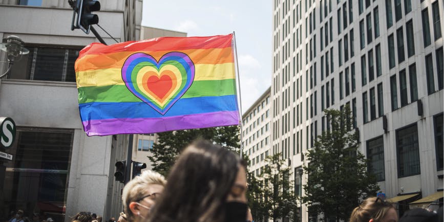 An outdoor photograph of city buildings and the heads of people marching in a Pride parade, with a pride flag with a heart at the center above the crowd.