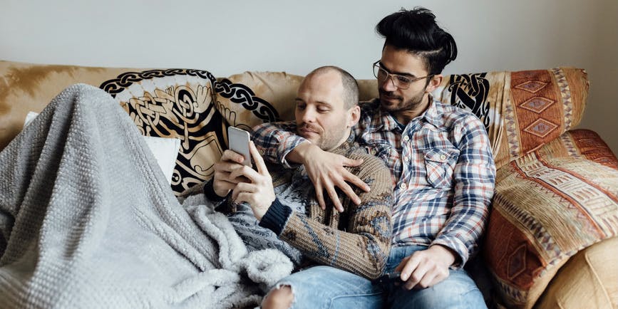 A color photo of two men—one with brown skin and hair and a mustache, wearing a plaid shirt, the other a white man with a shorn head wearing a brown sweater—cuddle on a couch under a blanket, looking at a phone.