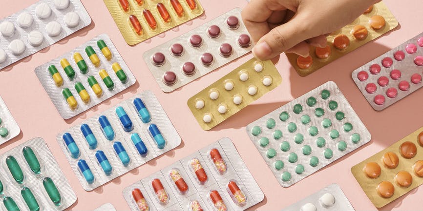 Different types of pills, Tablets in packs, and blister packs with colorful medicines are set on a pink background, with the fingers of a white hand picking one up.