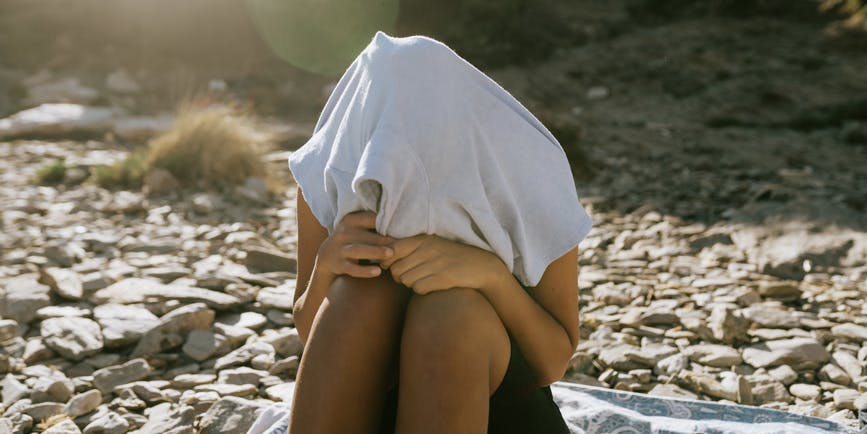 A young person with brown skin sitting on a beach blanket on a rocky beach has a t-shirt entirely covering their head.