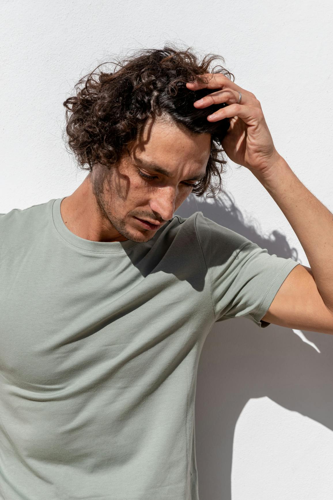 Un hombre de pie contra una pared blanca bajo el sol, pasando su mano por su cabello castaño y rizado mientras mira hacia abajo.