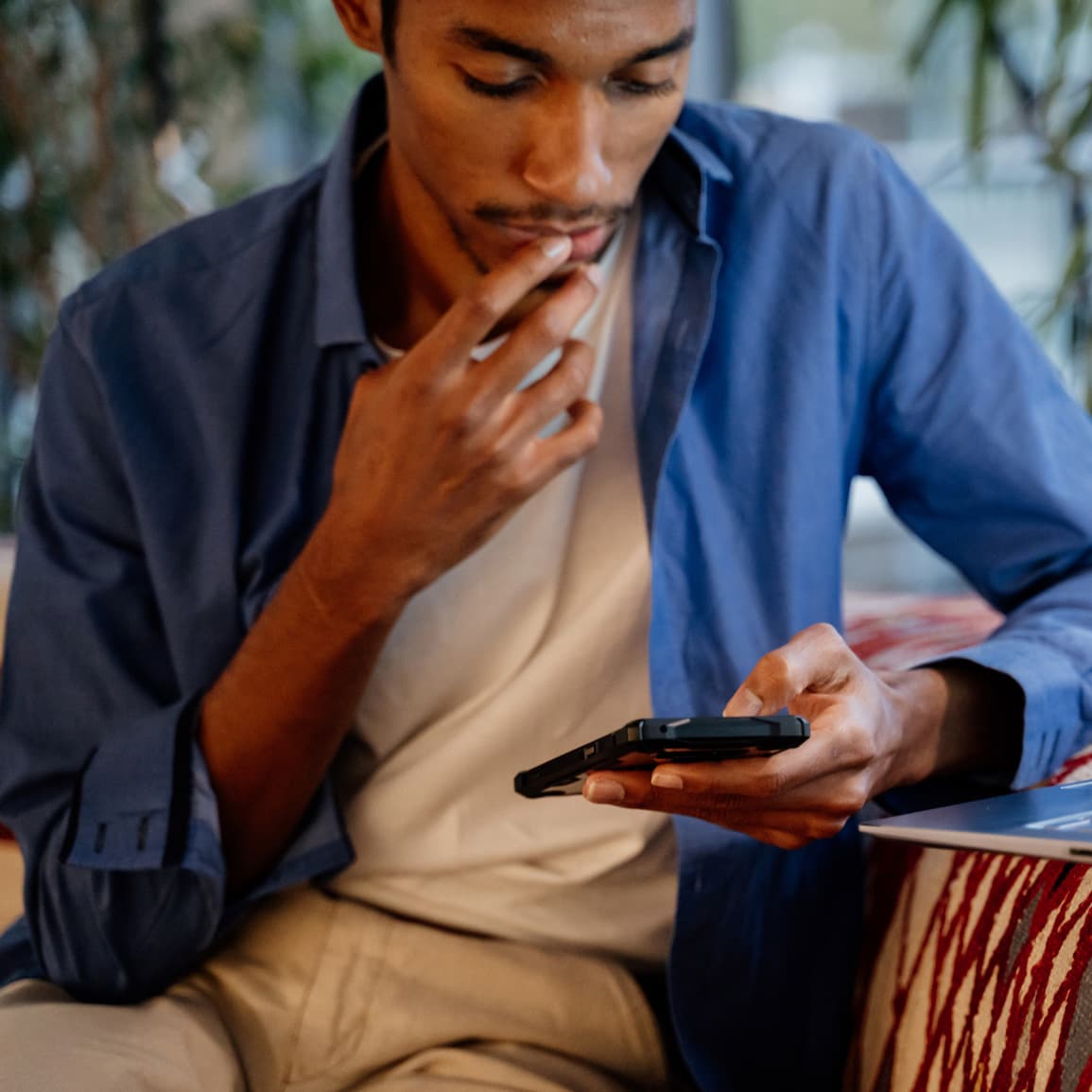 Young black man on phone, with a computer open in front of him, and touching his lip in thought