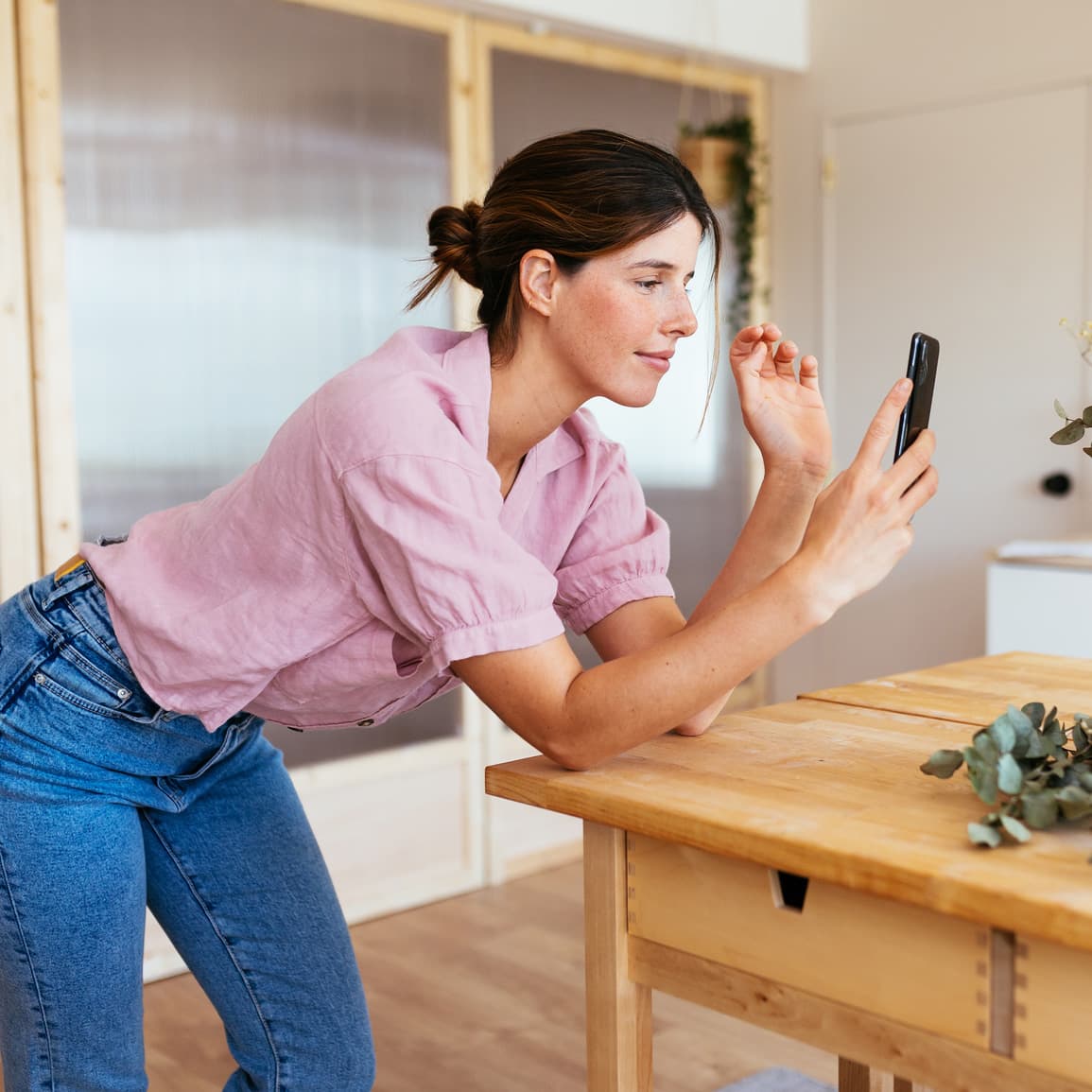 Mujer joven con una camisa rosa, inclinada sobre una isla de cocina, mirando y sonriendo levemente a su teléfono