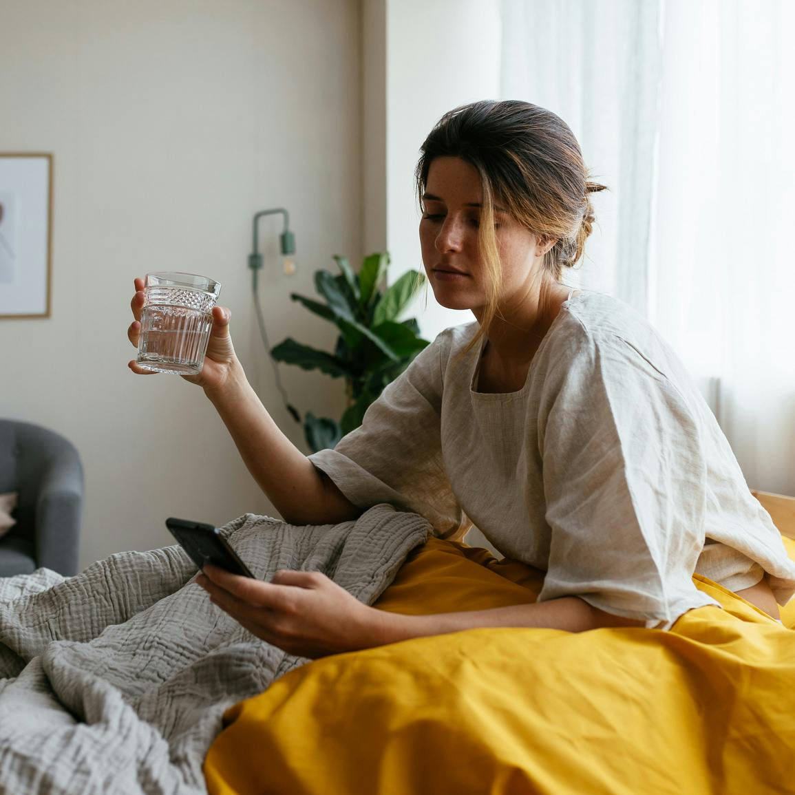 Mujer joven enferma en cama, sentada, debajo de mantas pesadas, con un vaso de agua en una mano y su teléfono en la otra, mirando el teléfono