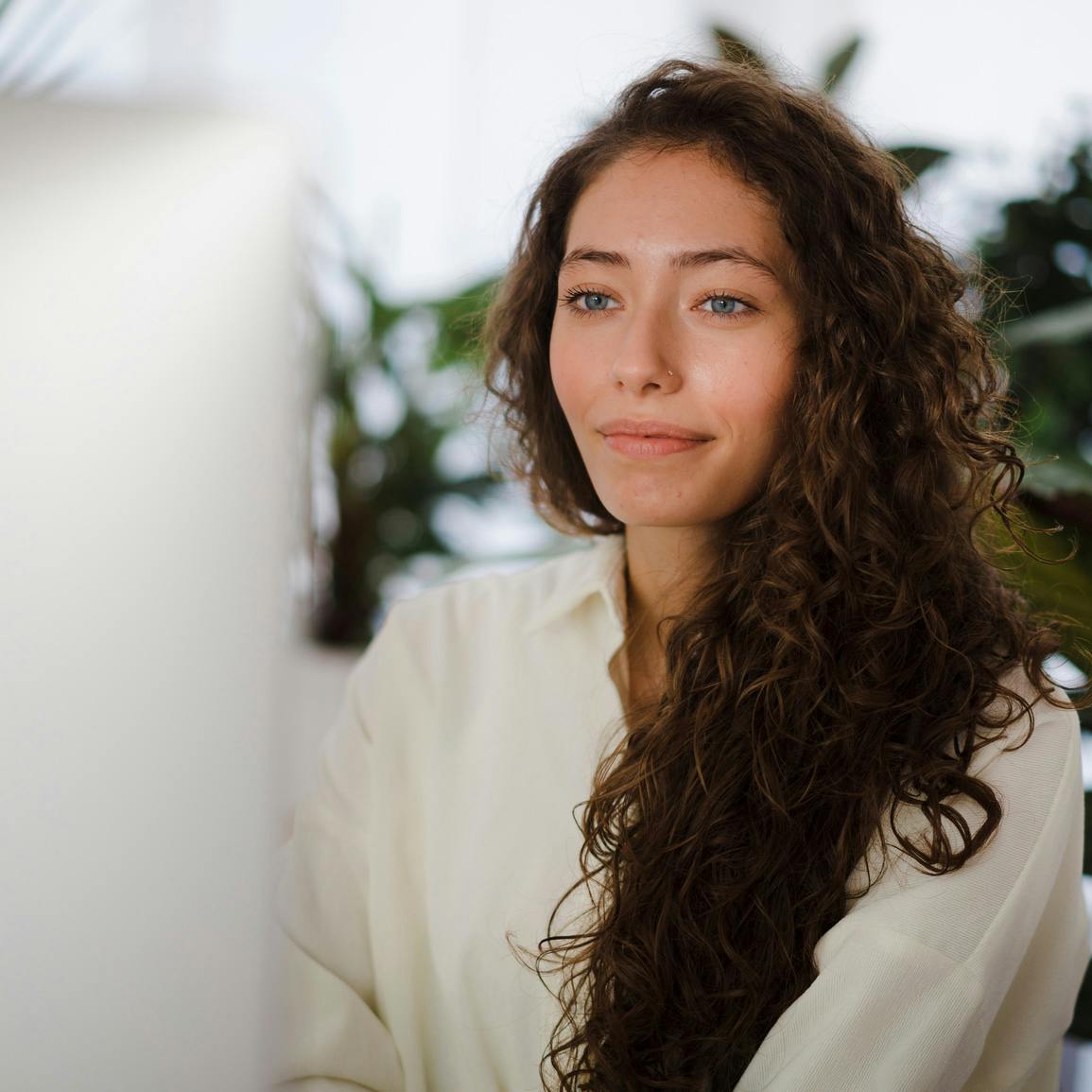 Woman with long dark curly hair and in white dress shirt sitting and smiling subtly at computer