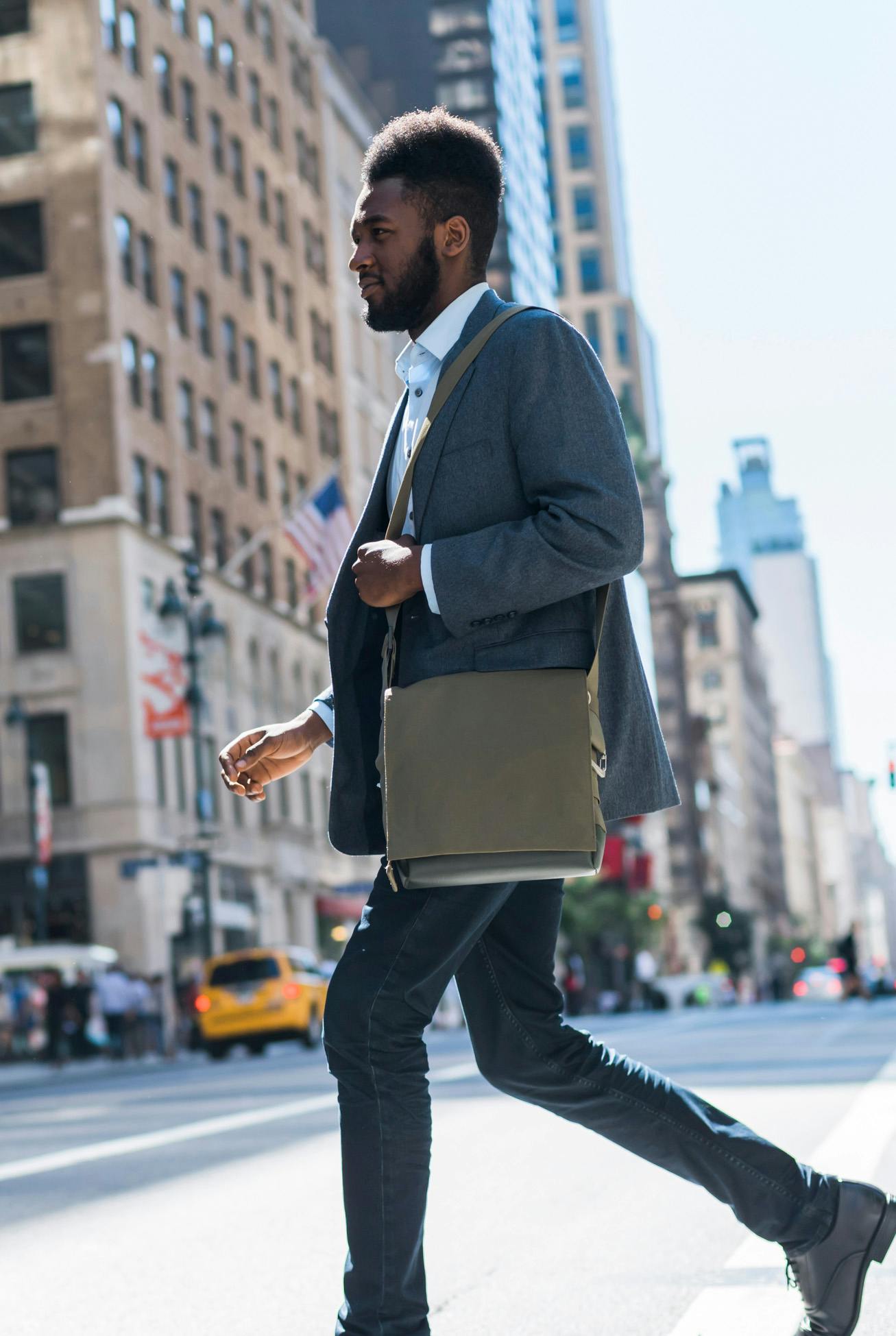 Hombre negro con chaqueta deportiva y jeans con bolsa de trabajo en el hombro, caminando con confianza por una calle de Manhattan