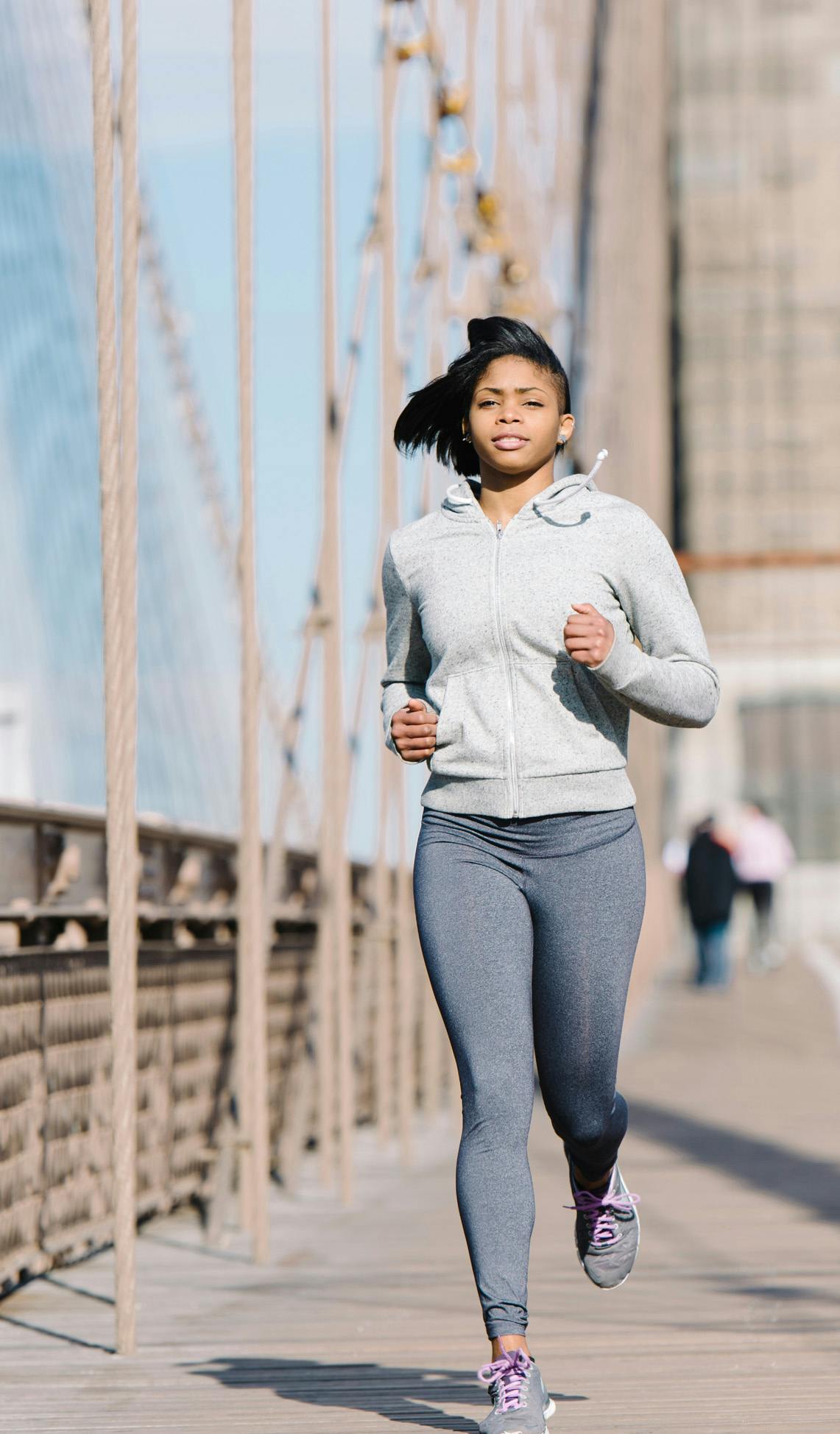 Black woman in heather gray hooded jacket running on the Brooklyn Bridge towards camera