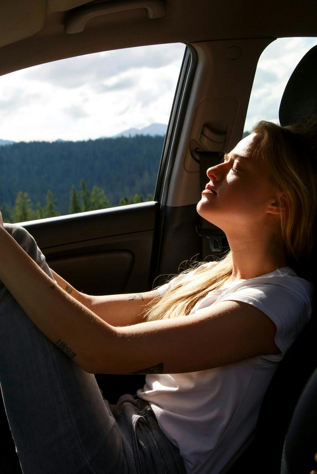 Woman in passenger seat of car with her head tilted back eyes closed. Sun is streaming onto her face and body. Outside the opened car window are mountainous forests.