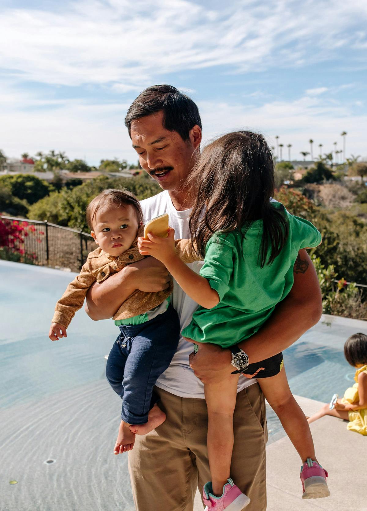 Padre asiático de unos cuarenta años con sus dos hijos pequeños en brazos, mientras camina junto a una piscina y sonríe