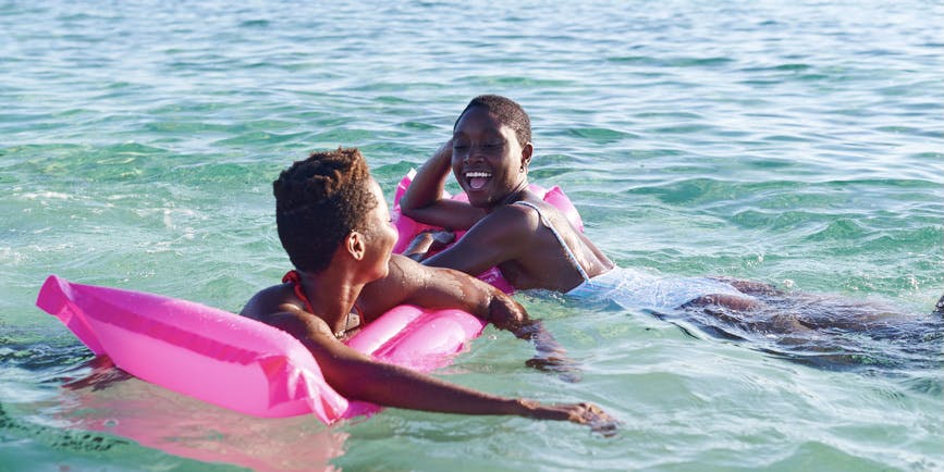 Dos jóvenes amigas africanas que ríen se aferran a una carroza mientras nadan juntas en el océano en un día soleado.