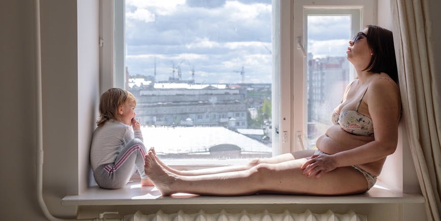 Curvy white woman dressed in a bikini sunbathes inside on a windowsill while their toddler looks out the window.