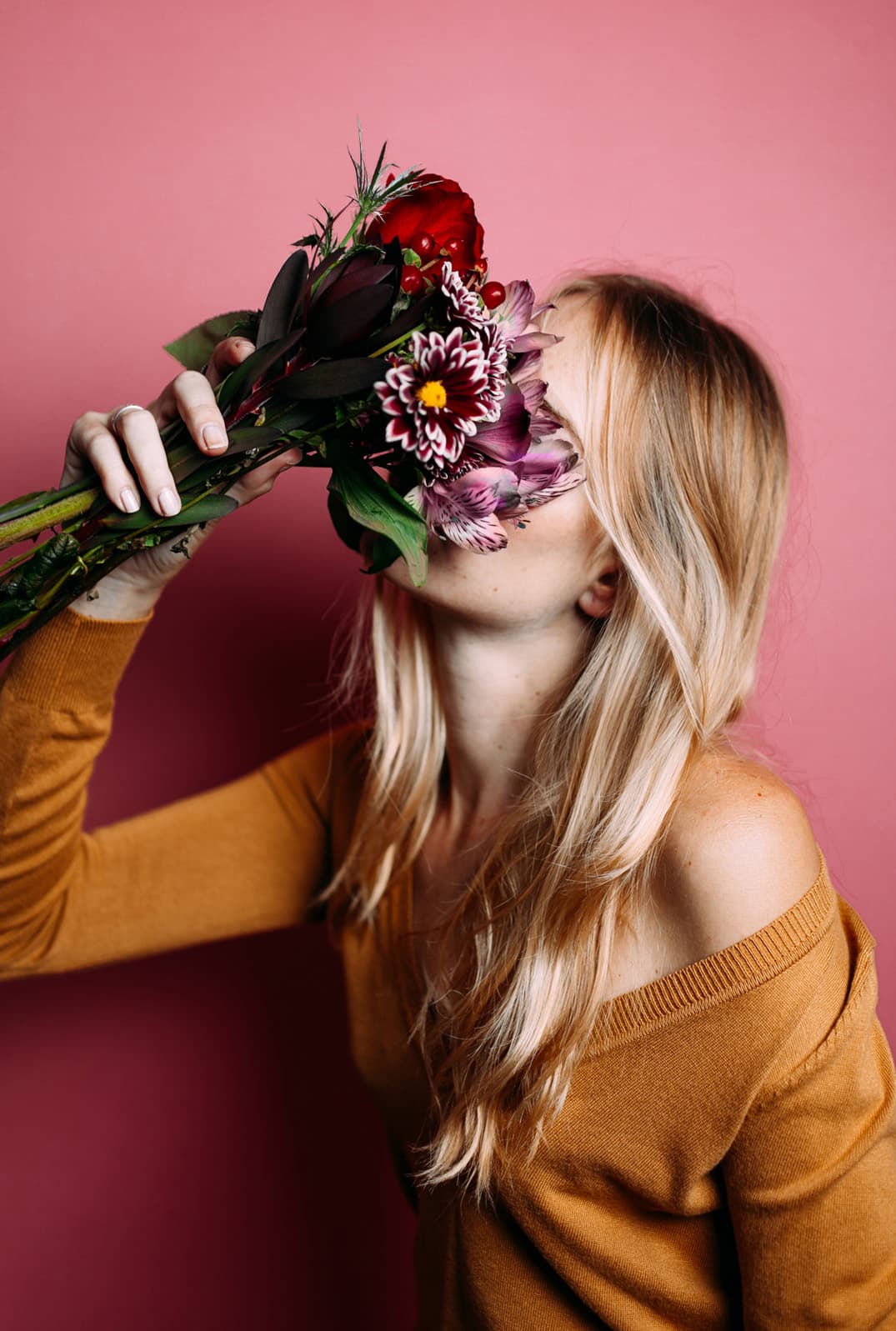 Una mujer rubia enterrando su rostro en un ramo de flores, de pie contra una pared roja