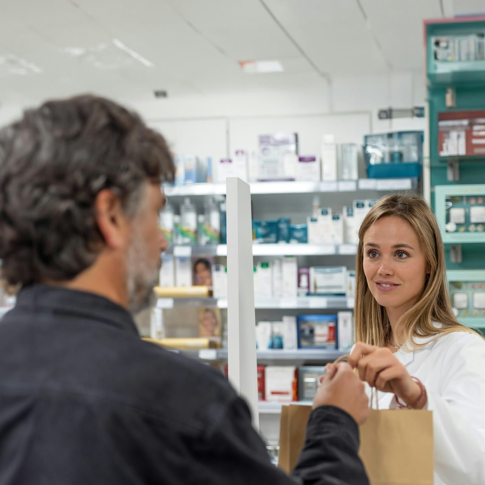 Un hombre canoso recoge una receta de un farmacéutico sonriente, quien le entrega una bolsa en el mostrador de la farmacia.