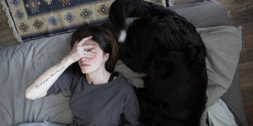 Young white woman with brown hair wearing a gray t-shirt lays on the floor next to a large black dog and covers her eyes with her hand.