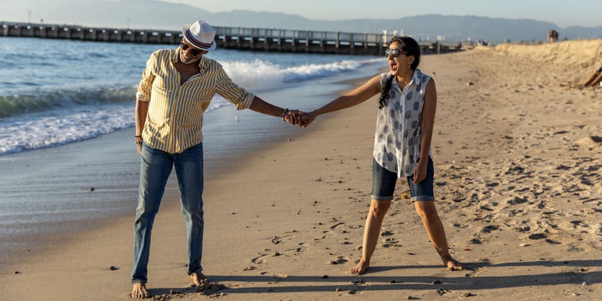A senior couple with brown/tan skin wearing jeans and summer shirts holds hands while walking on the beach, smiling at each other.  