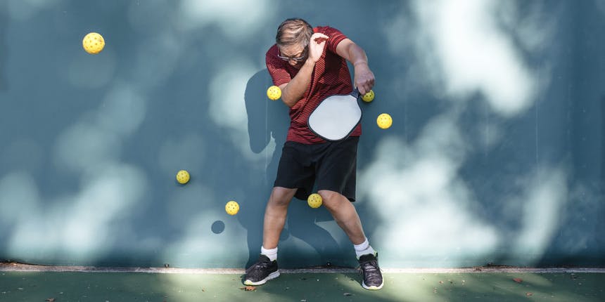 A white man wearing a red shirt and black shorts stands against a blue wall holding a pickleball racket while yellow balls fly at him.