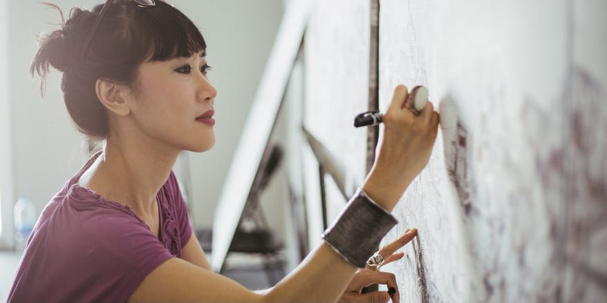 Una mujer asiática vestida con una camisa morada y el cabello castaño recogido se concentra en dibujar en su lienzo con un bolígrafo. La artista trabaja en un estudio con luz natural.