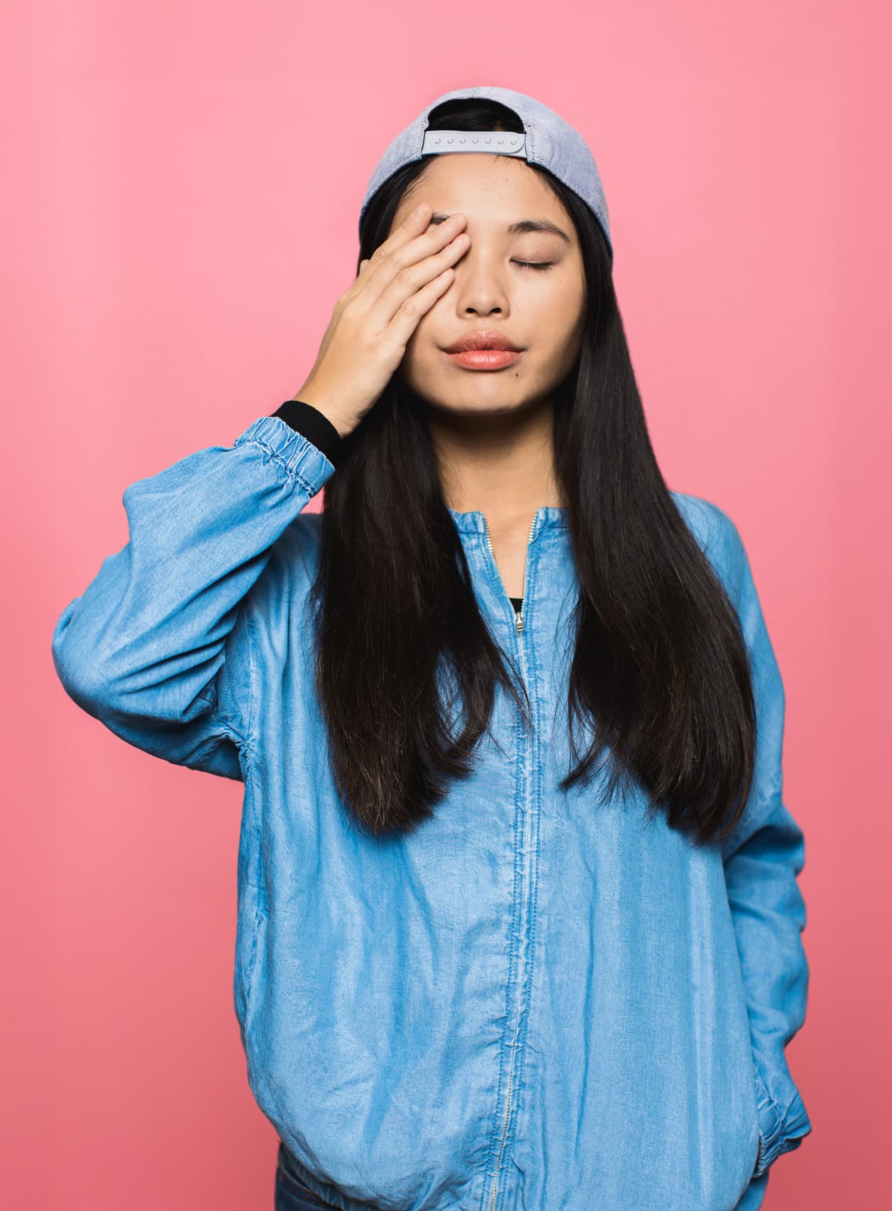 Mujer asiática con chaqueta azul y gorra de béisbol azul claro con los ojos cerrados al revés con la mano sobre un ojo.