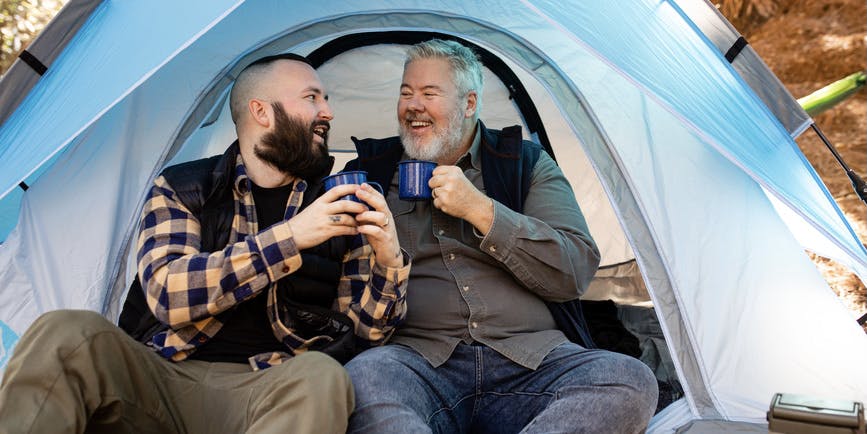 A happy LGBT couple laugh together in their blue tent while holding blue camping mugs at their campsite.