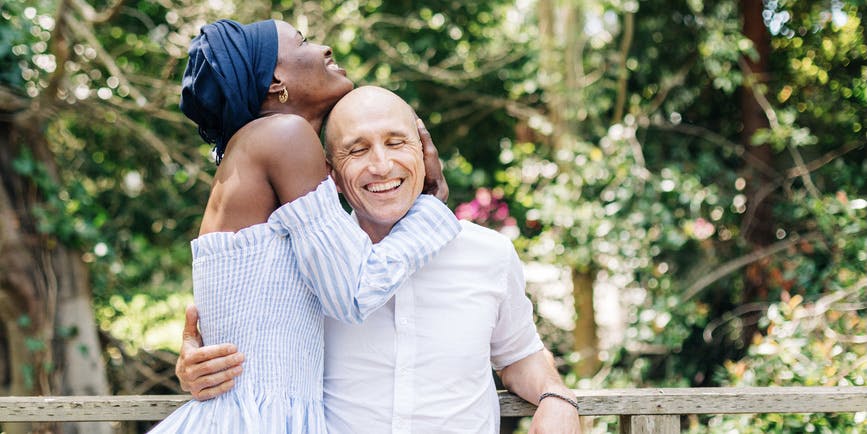 Happy mature white man smiles as he's embraced by a gorgeous young Black lady in a blue turban standing on a wooden balcony in nature.