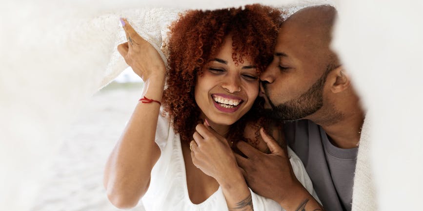 Una joven pareja dominicana se abraza y besa tiernamente bajo una manta blanca.