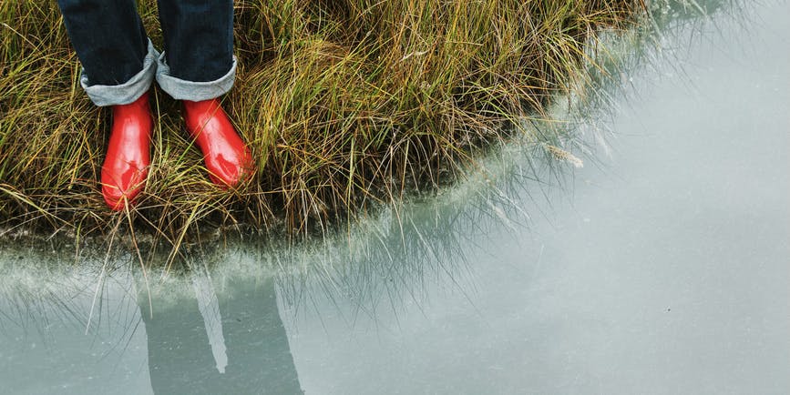 An overhead shot of the legs of a person wearing dark blue jeans and red rubber boots standing in the grass at the edge of still misty-blue water.