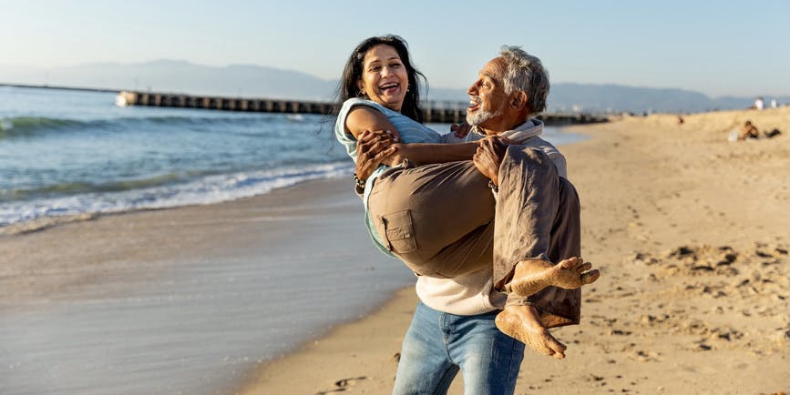 A senior couple with brown skin wearing long pants and t-shirts is on an empty beach. The man holds the woman in his arms, and both are smiling.