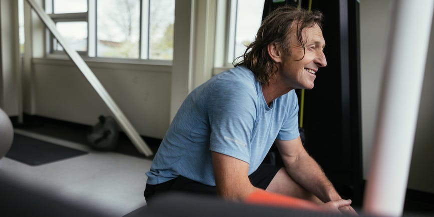 A white man with long hair in his fifties wearing a blue t-shirt smiles as he sits on a piece of gym equipment.