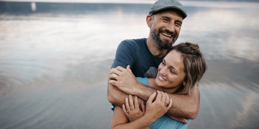 Una joven pareja blanca con camisetas se abraza en una playa con aguas tranquilas a sus espaldas. Ambos sonríen y se abrazan con fuerza.