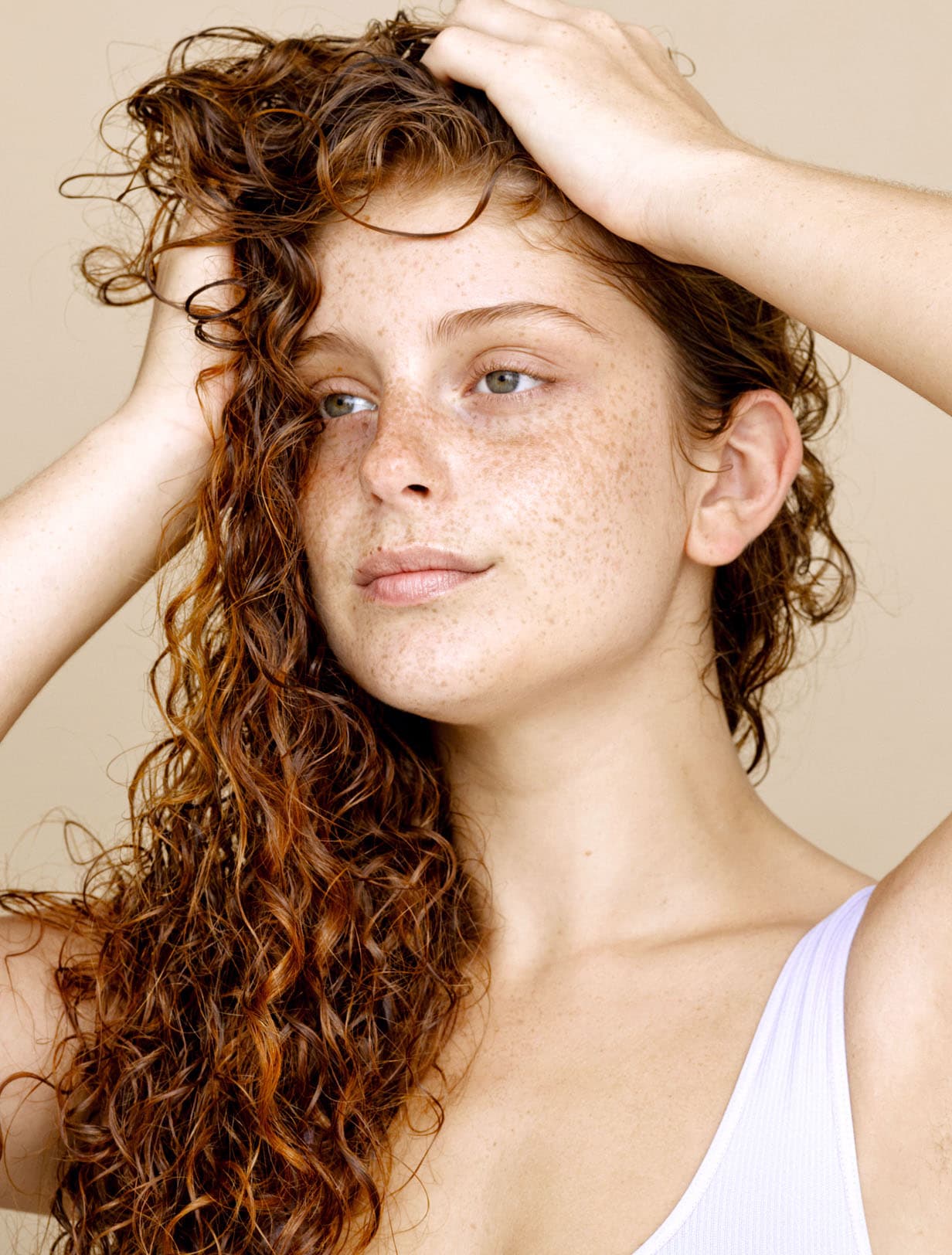 Mujer con cabello rojo rizado y pecas marcadas que sostiene su cabello con ambas manos, mirando a lo lejos