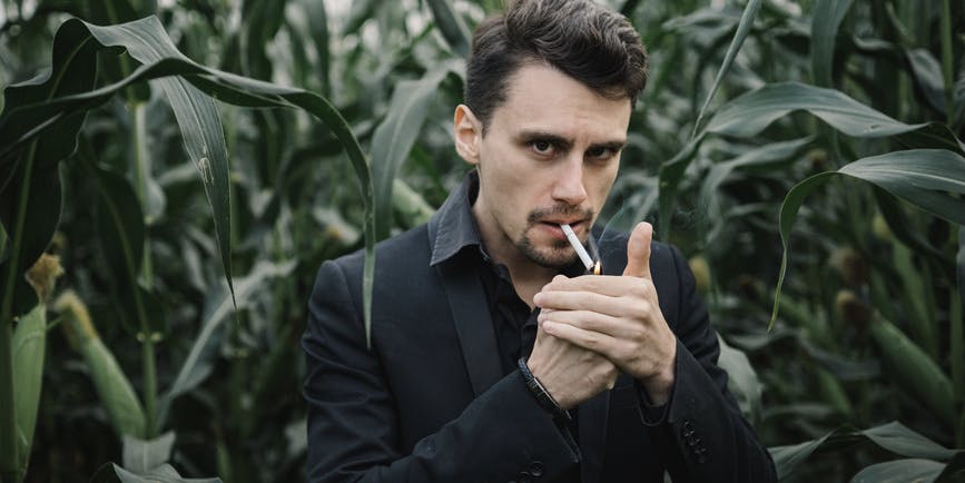 A young white man with short brown hair dressed in black suit stands in a corn field and looks at the camera as he lights a cigarette.