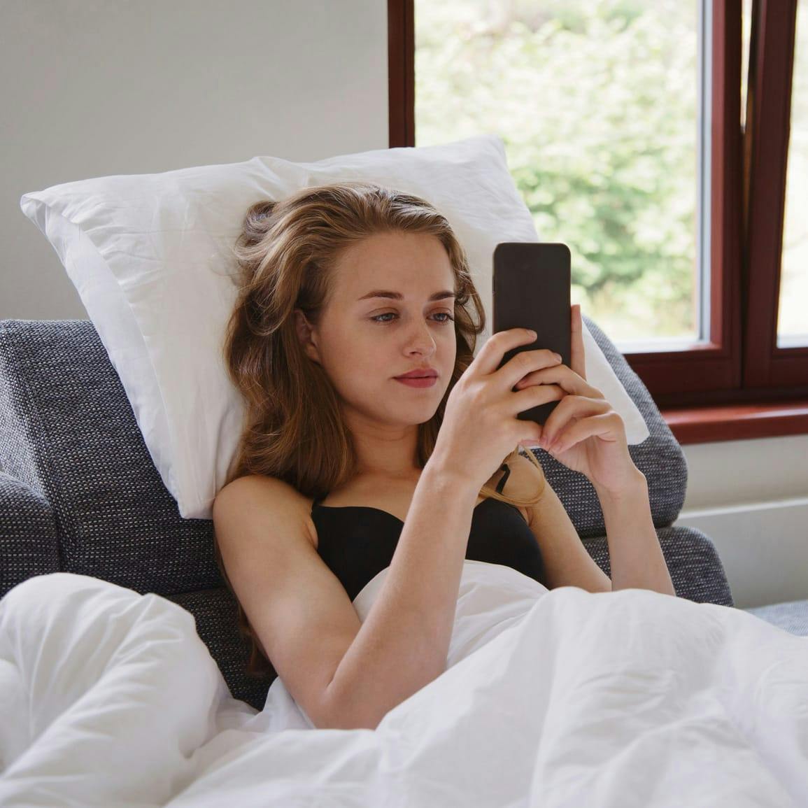 Woman sitting in up bed, covered in sheets, both hands on her phone