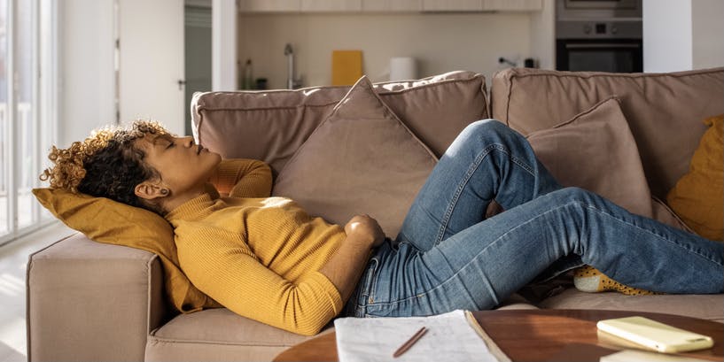 Side view of calm Hispanic woman in casual clothes closing eyes and resting on couch near table with smartphone and notebook while taking break in home studies in light living room.
