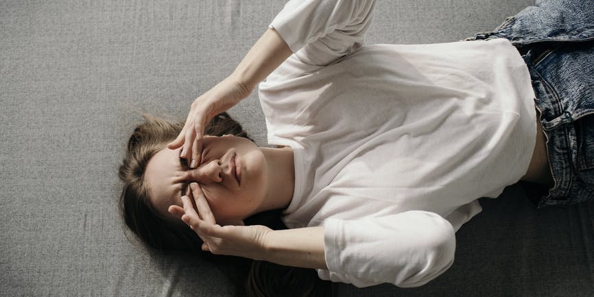 From above, a young white woman lays on her bed wearing a white shirt and jeans, frowning as she rubs her face in pain or fatigue.
