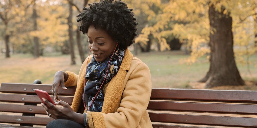 Una mujer negra con un abrigo naranja se sienta en un banco del parque con árboles otoñales detrás de ella y sonríe mientras mira su teléfono.