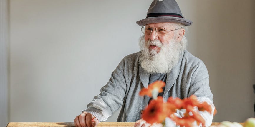 Enchanting grey-bearded white senior man with an elegant style looks away from the camera and smiles while sitting at a kitchen table at home.