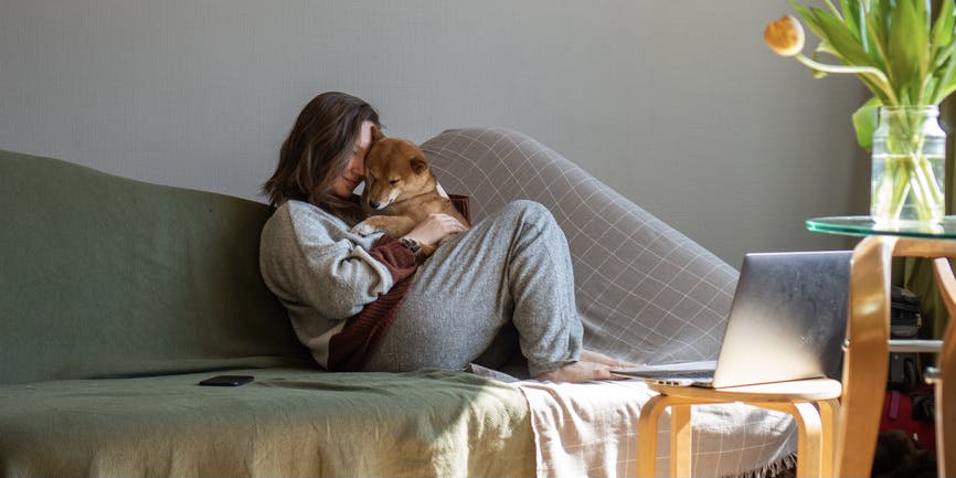 A curvy white woman wearing sweatpants cuddles with her shiba inu on her living room couch.