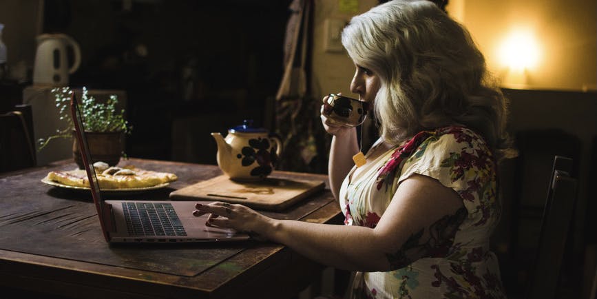 Beautiful curvy woman with grey hair working on her laptop while drinking tea while a candle burns behind her.