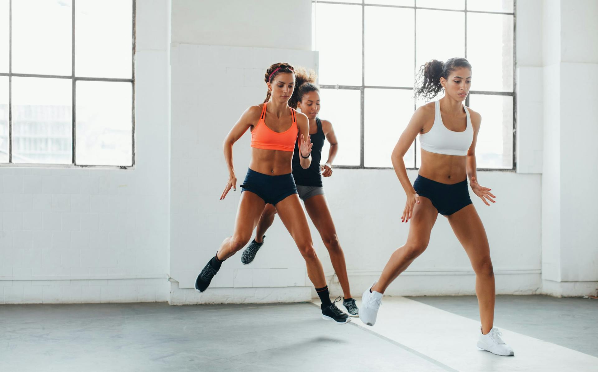 Three women wearing sports bras and shorts in a fitness class in a gym with bare walls and large windows jumping from foot to foot