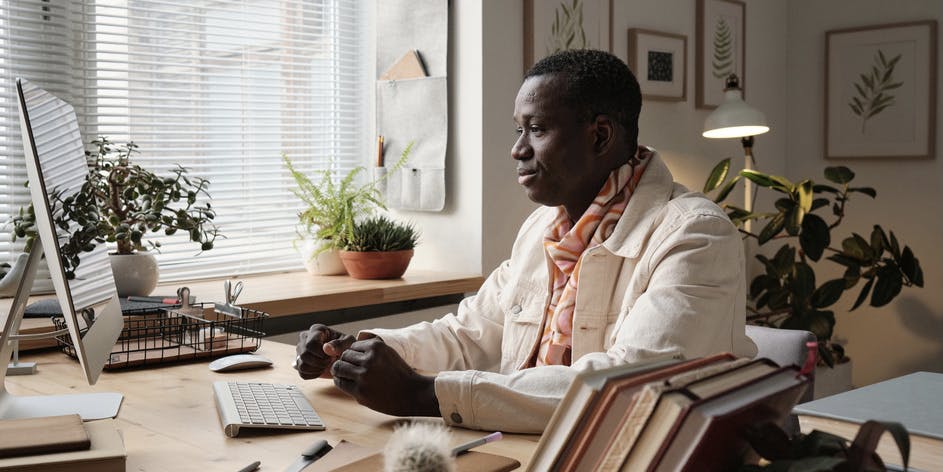 Horizontal medium portrait of handsome young African American man wearing fashionable clothes sitting at desk at home looking at computer screen.