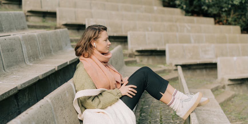 Woman sitting alone in a relaxed position in outdoor amphitheatre. She is listening to music using earbud headphones and wearing warm clothes.
