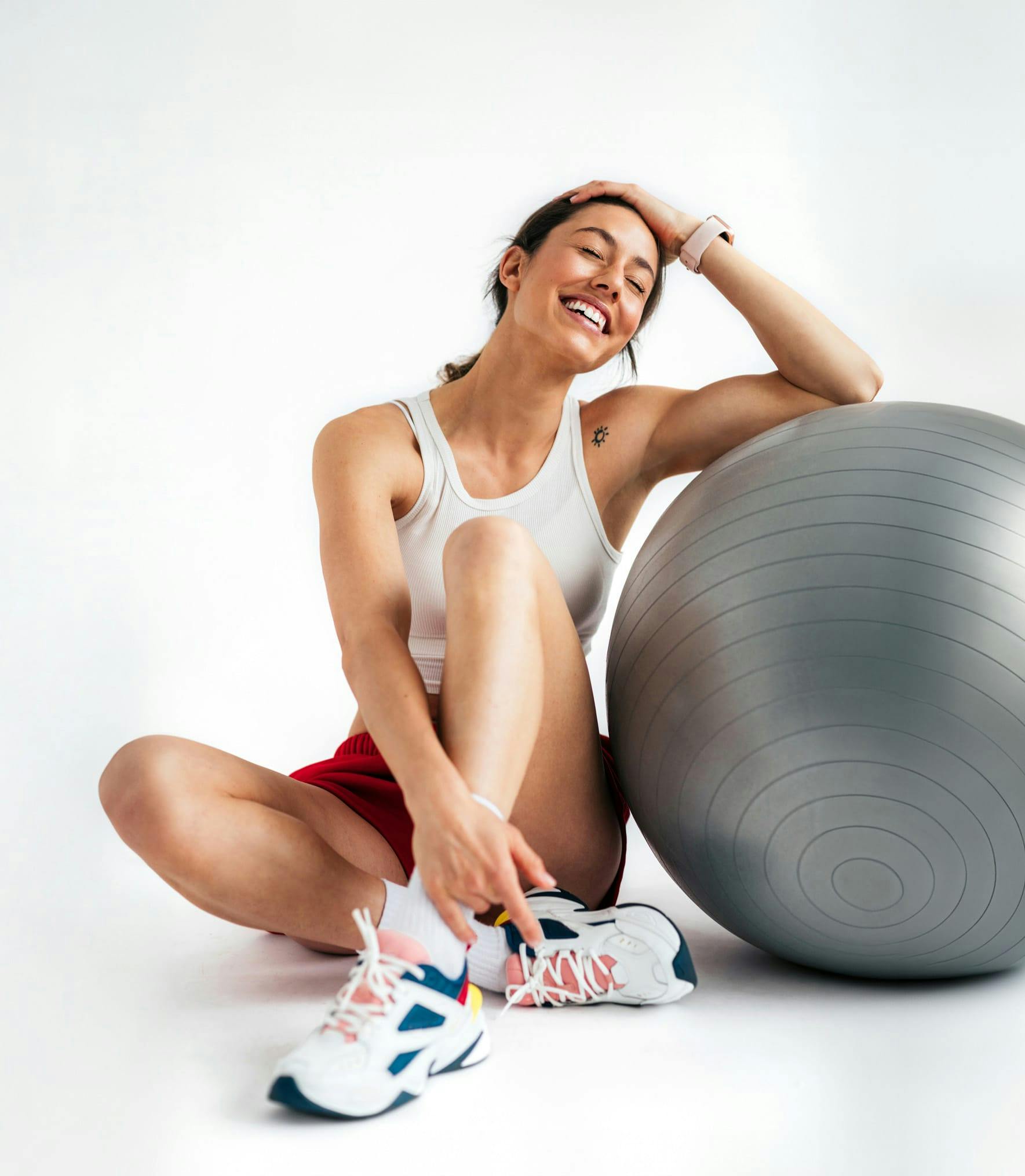 A woman in workout clothes sitting and smiling leaning into a workout ball