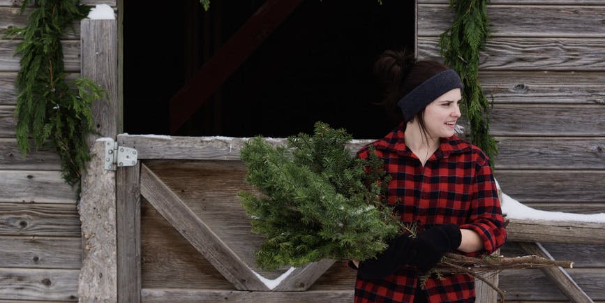 A young white woman wearing a black and red plaid shirt stands in front of a barn holding boughs of evergreens.
