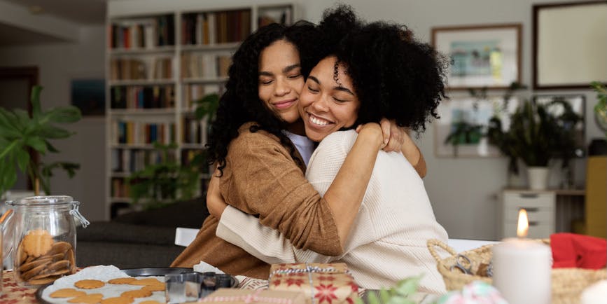Close-up of two young happy Black women embracing while preparing xmas cookie boxes at home.