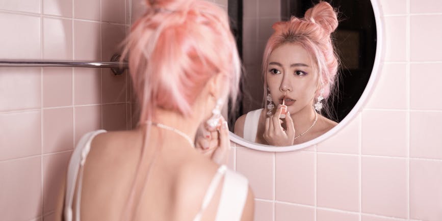 An Asian woman with pink hair puts lipstick on while looking in the mirror of a pink bathroom.