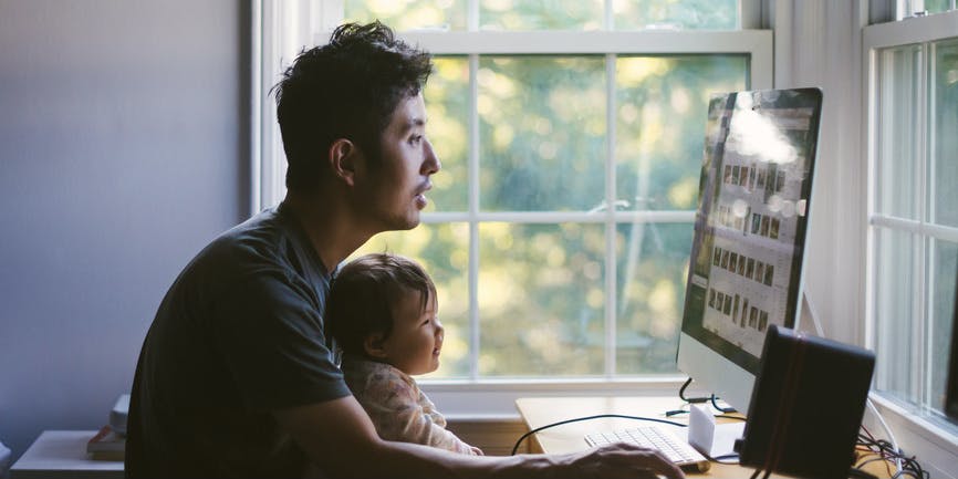 A young Asian dad works on his computer at home with baby on his lap, smiling and looking at screen together.