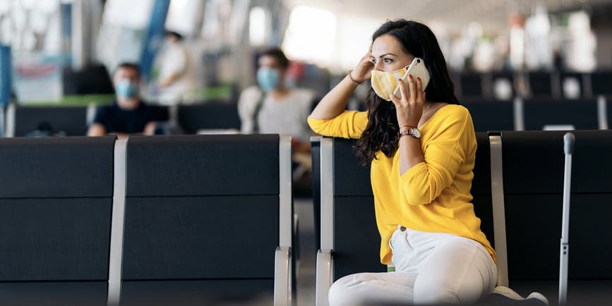Photo of middle aged white woman wearing a yellow sweater and a face mask, using her phone while she waits for her flight in an airport.