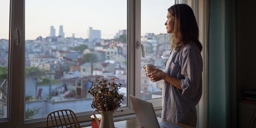 Una joven blanca de cabello largo y castaño mira fijamente la puesta de sol menguante mientras sostiene una taza de café, con un portátil abierto sobre la mesa de al lado.