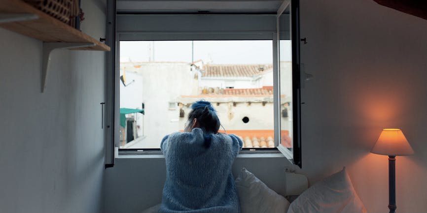 A young white woman in warm pajamas sits on the bed of her cozy house, looking out the window with her back to the camera while in a dim room.
