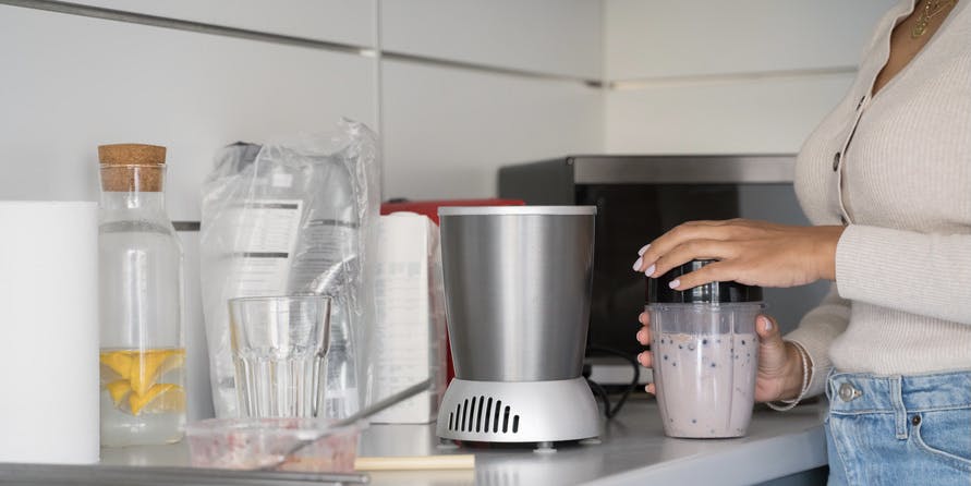 Close up photo of a woman with brown skin wearing a tan shirt preparing a breakfast smoothie. 